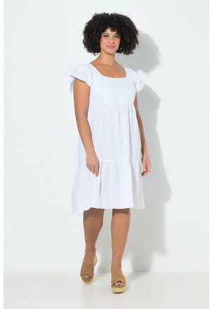 Kiharatukkainen nainen, jolla on valkoinen polvipituinen mekko, jossa liehuvat hihat, ja beigen väriset kiilakorkokengät, seisoo valkoista taustaa vasten.