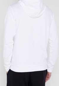 Sweat à capuche blanc avec une texture douce, capuche attachée, manches longues et ourlet côtelé. Vue de dos montrant un design simple, sans logos ni motifs visibles.