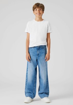T-shirt en coton blanc léger associé à un pantalon en denim bleu à jambes larges et amples, avec un délavage et des poches avant. Baskets blanches.