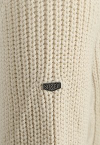 Dzianinowy kremowy sweter o prążkowanej strukturze, z czarną metalową metką z wytłoczonym napisem "SD&CO" z boku.