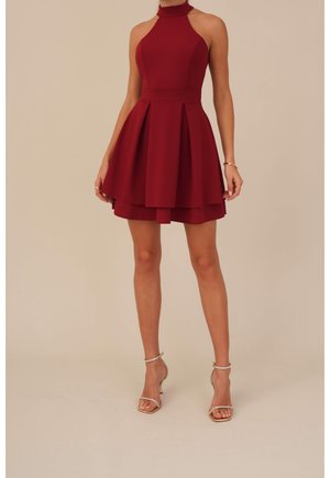 Frau in ärmellosem burgunderrotem Neckholderkleid mit plissiertem Rock und goldfarbenen Sandaletten mit hohem Absatz vor schlichthintergrund in Beige.