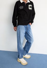 Polo negra de manga larga con cuello blanco, con texto y logo bordados; combinada con jeans azul claro y zapatillas beige.