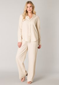 Beiges Leinenhemd mit Knöpfen, langen Ärmeln und einer Brusttasche, kombiniert mit locker sitzenden beigen Leinenhosen und Riemchenabsatzschuhen.