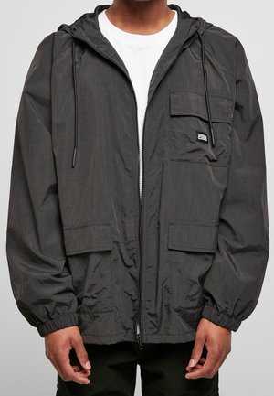 Personne portant une veste noire à capuche avec fermeture éclair, poignets élastiques et plusieurs poches à rabat sur une chemise blanche.
