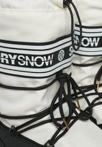 Bottes de neige blanches avec une semelle noire épaisse, ornées de lacets noirs et d'une bande de logo bien visible. La tige présente une finition en tissu texturé.
