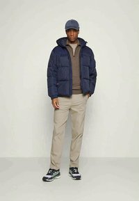 Marineblå puffjakke med hette, beige genser, beige bukser og sorte joggesko med grønne detaljer. Bakgrunnen er i en lys, nøytral farge.