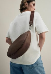 Braune Lederhandtasche mit einer weichen, runden Form und silberner Hardware, crossbody über einem weißen lockeren Hemd und blauen Denim-Shorts getragen.