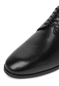 Fekete bőrcipő sima felülettel, hegyes orral és díszes fűzéssel. Erős talppal és diszkrét varrással rendelkezik.