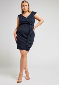 Meriino sinine ümbrikkleit sügava V-dekoltee, laiade õlavarrede ja volangiga külje detailiga. Sile tekstuur, keha lähedane stiil, koos kuldsete sandaalidega.