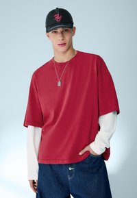 Mladý muž s černou čepicí, červeným oversized tričkem přes bílými dlouhými rukávy, volnými modrými džínami a stříbrným přívěskem na krku, stojící na jednobarevném pozadí.