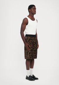 Homme portant un débardeur blanc sans manches, un short genou marron et noir à motifs, des chaussettes blanches et des mocassins noirs, se tenant devant un fond blanc.
