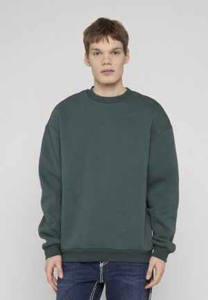 Młody mężczyzna z krótkimi brązowymi włosami, ubrany w ciemnozieloną, oversize'ową bluzę i niebieskie dżinsy, stoi na tle jasnego, jednolitego tła.