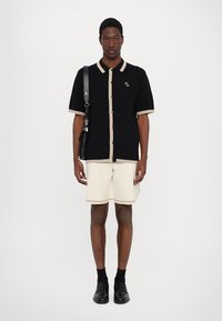 Schwarzes Polo mit kurzen Ärmeln und beigen Akzenten sowie Knöpfen, kombiniert mit beigen Shorts mit passendem Besatz. Das Modell trägt eine schwarze Umhängetasche.