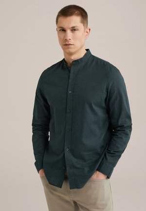 Camisa de manga larga verde oscuro con cuello mao, botones en la parte delantera y tejido texturizado; el modelo lleva pantalones beige claro, con las manos en los bolsillos.