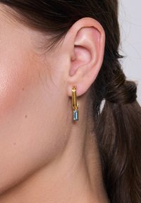 Złota kolczyk w kształcie koła z prostokątnym wisiorkiem z niebieskiego kamienia szlachetnego, prezentujący gładkie tekstury i polerowane wykończenie.