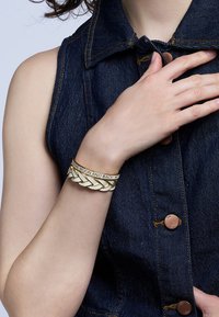Bracelet doré avec un design tressé et un message grav�é. Porté au poignet, créant un contraste avec un haut en denim foncé.