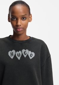 Juodas džemperis su apvalia iškirpte ir laisvo silueto. Priekinėje dalyje dizainas su žodžiu „HUGO“ balta spalva, apvyniotas pilkų širdelių kontūrais.