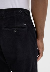 Black corduroy pants featuring a textured ribbed design, back pockets with buttons, and a small brand label near the waistband.