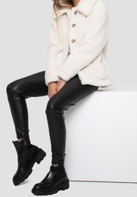 Veste en fausse fourrure crème avec un col et quatre boutons, associée à un pantalon en fausse cuir noir et des bottines noires épaisses.