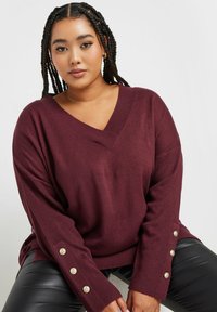 Burgunderfarbener Strickpullover mit V-Ausschnitt, lässiger oversize Passform und langen Ärmeln, die mit goldenen Knopfakzenten an den Bündchen versehen sind. Glatte Textur, lässiges Design.