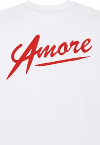Balts kokvilnas T krekls ar sarkanu slīprakstu tekstu "Amore" un plašu apakšsvītru, demonstrējot maigu tekstūru un neformālu dizainu.