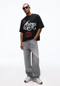 Fekete, túlméretezett póló fehér és piros grafikus szöveggel; szürke farmerrel párosítva; fekete napszemüveg; világos tornacipő szürke részletekkel.