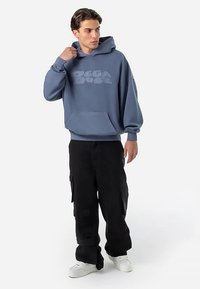 Νεαρός άνδρας που ρυθμίζει την κουκούλα μπλε oversized φούτερ με το κείμενο "DECADOGS", φορώντας μαύρο cargo παντελόνι και άσπρα αθλητικά παπούτσια, στέκεται σε άσπρο φόντο.