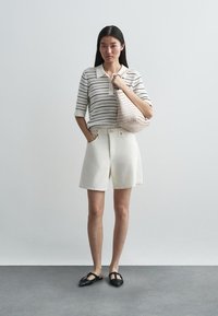 Krämfärgad stickad polo med svarta ränder, vita shorts och svarta platta skor. Bär en ljusfärgad vävd väska. Enkel design, avslappnad passform.