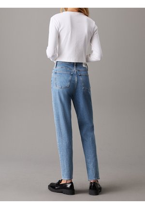 Femme portant un jean taille haute bleu clair, un crop top blanc à manches longues et des mocassins noirs, debout de dos sur un fond gris.