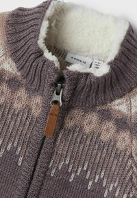 Sweter z dzianiny na zamek błyskawiczny z brązowym, kremowym i beżowym wzorem, ściągaczem przy kołnierzu, skórzaną zawieszką zamka oraz miękką białą polarową podszewką.