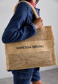 Geweven stro tas met een rechthoekige vorm en platte bodem. Voorzien van zwarte tekst "VANESSA BRUNO" en twee stevige, gestructureerde handvatten.