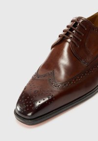 Zapato de vestir de cuero marrón con puntera afilada, decoración de broguing y un acabado suave. Presenta cordones y una suela negra contrastante.