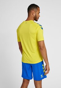 Camisa activa amarilla de manga corta, con acentos en chevron azul en los hombros, combinada con pantalones cortos azules. Textura suave.