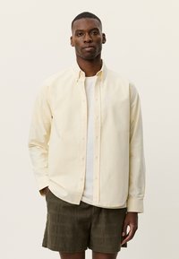 Ανοιχτό κίτρινο βαμβακερό πουκάμισο με κουμπιά, με μακριά μανίκια και χαλαρή εφαρμογή, φορεμένο πάνω από λευκό T-shirt και συνδυασμένο με σορτς με σχέδια.