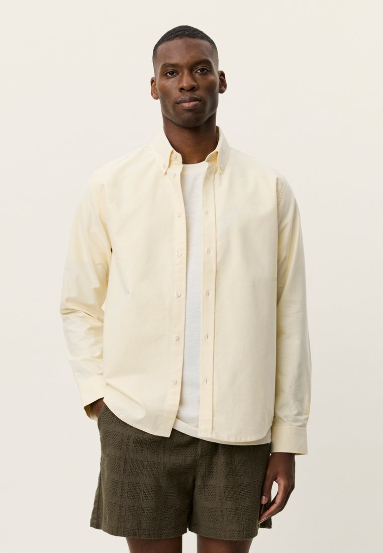 Ανοιχτό κίτρινο βαμβακερό πουκάμισο με κουμπιά, με μακριά μανίκια και χαλαρή εφαρμογή, φορεμένο πάνω από λευκό T-shirt και συνδυασμένο με σορτς με σχέδια.