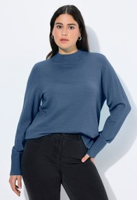 Modrý pletený svetr s vysokým límcem a dlouhými rukávy, se žebrovanými manžetami a volným střihem, který je kombinován s černými džíny.