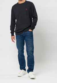 Mörkgrå crew-neck tröja med ribbade manschetter, i kombination med blå jeans och vita sneakers. Har en liten röd logga på bröstet.