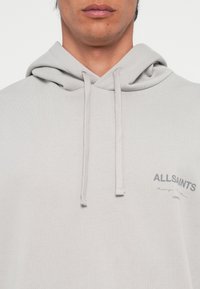 Haut à capuche gris clair en coton doux, avec un cordon de serrage, un logo sur le devant en gris foncé et des poignets côtelés. Texture lisse avec un design minimaliste.