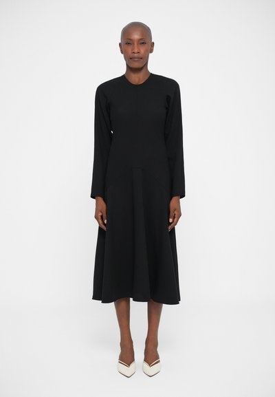 Woman with shaved head wearing a long-sleeve black midi dress and white pointed-toe heels, standing against a plain white background.