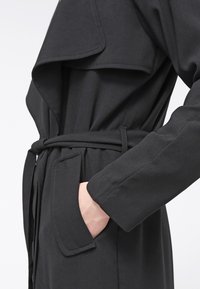 Černý trench coat s páskem, širokými klopami, postranními kapsami a dlouhými rukávy. Hladká struktura látky s jemnými detaily šití.