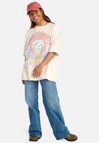 Beige oversized t-shirt with colorful graphic design featuring waves and flowers. Paired with light blue wide-leg jeans and brown shoes. Pink cap.
