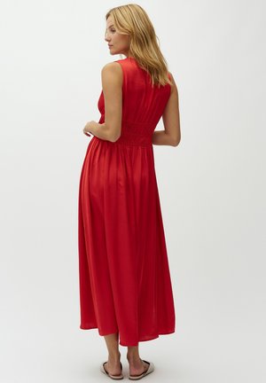 Blondidega naine, kes kannab varrukateta punast midi-kleiti kogutud vöökohaga, seisab paljajalu valge tausta ees.