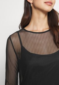 Femme portant un haut noir transparent à manches longues avec des rayures verticales par-dessus un caraco noir uni, montrant son cou et une partie de son visage.