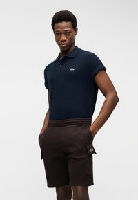 Άνδρας που φοράει μπλε ναυτικό πόλο μπλουζάκι και σκούρα καφέ cargo σορτς με τα χέρια στις τσέπες, μπροστά σε απλό φόντο.