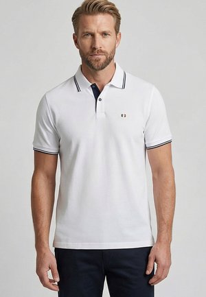 Man met baard die een wit poloshirt met korte mouwen draagt met marineblauwe bies aan kraag en mouwen, staand tegen een effen achtergrond.