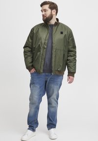 Maslinasto zelena bomber jakna s patentnim zatvaračem sprijeda, bočnim džepovima i rebrastim završecima. Nošena preko tamne veste, u kombinaciji s plavim trapericama i bijelim tenisicama.