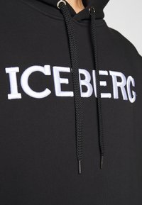 Gros plan d'un sweat à capuche noir avec de grosses cordelettes noires et le logo brodé "ICEBERG" en blanc sur la poitrine.