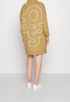 Person iført en oversized sandfarvet hættetrøjekjole med abstrakt hvid linjedesign på ryggen, kombineret med hvide sneakers, stående med ryggen mod.
