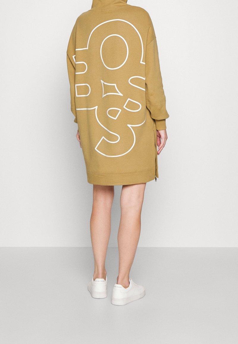 Personne portant une robe pull oversize beige avec un motif abstrait de lignes blanches au dos, associée à des baskets blanches, debout, de dos.