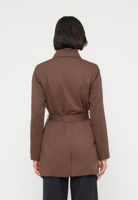 Καφέ παλτό με ζώνη και κομψό σχέδιο, μακριά μανίκια και κλασικό γιακά. Το ύφασμα φαίνεται λείο με δομημένη σιλουέτα.